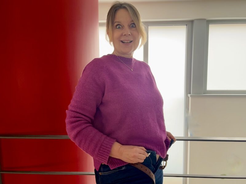 Frau blickt schockiert an sich herab, weil ihre Jeans am Bauch nicht mehr zugeht – ein typisches Symptom für den Hormonbauch